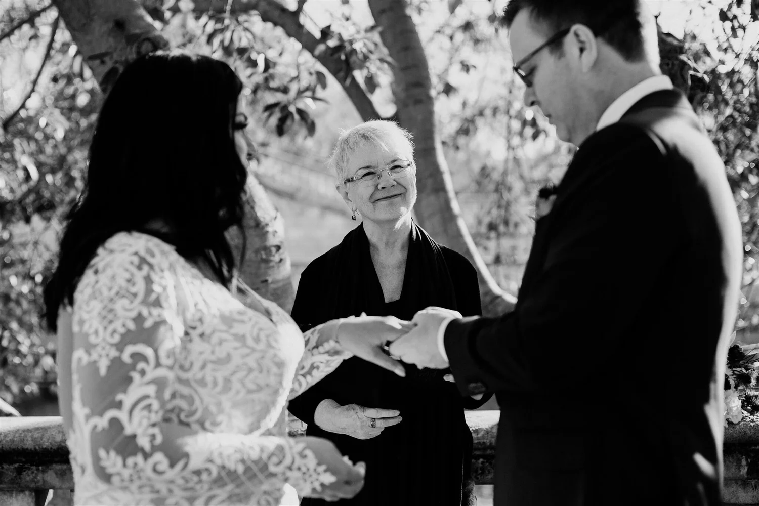 A black and white photo of a couple holding hands, facing each other, during their wedding ceremony outdoors. They are standing before an officiant who is smiling and looking at them. The couple is dressed in wedding attire, with the woman in a lace