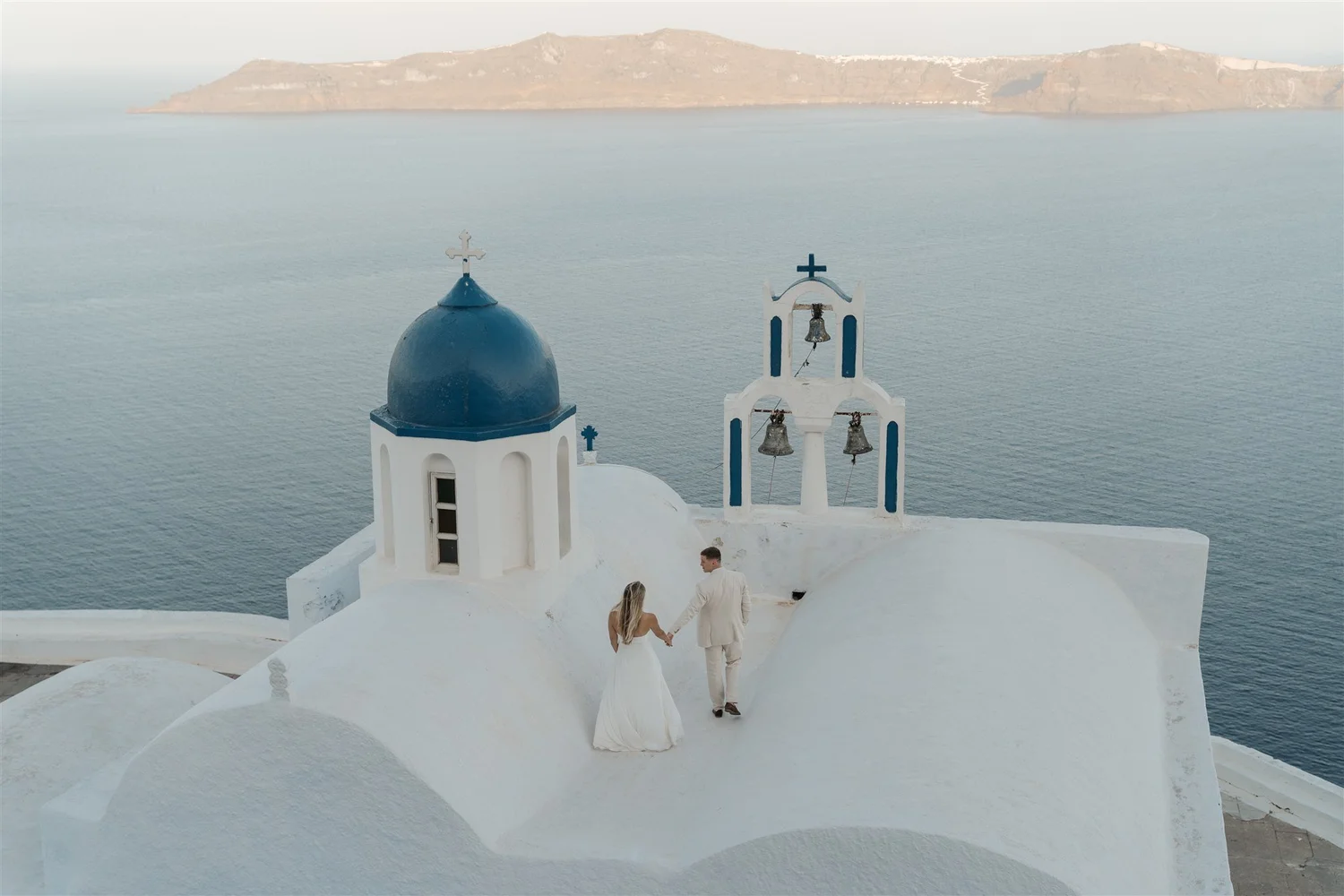 A bride and groom hold hands and walk along white-washed buildings with domed roofs and church bells, overlooking the sea in Santorini, Greece.