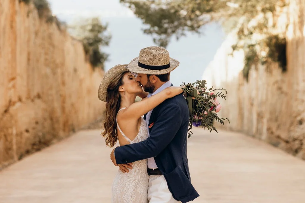 A couple sharing a kiss with their arms around each other on a cobblestone street in a warm outdoor setting, both wearing sun hats and the woman holding a bouquet of flowers.