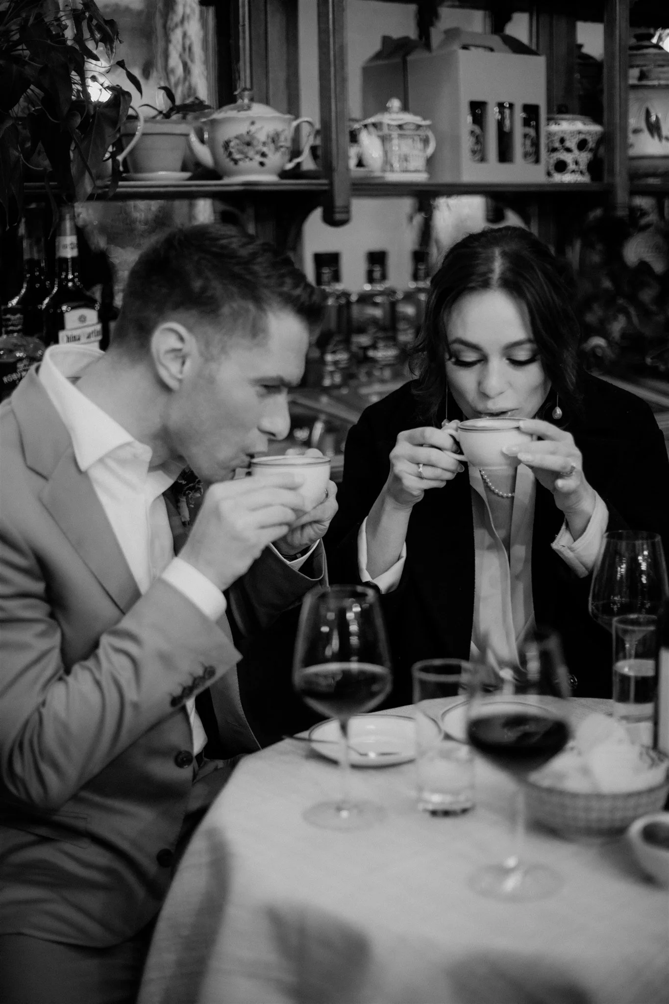 A black and white photo of two people, a man and a woman, sitting at a dining table and drinking from cups. The table has glasses and plates, and the background has shelves with china and bottles.