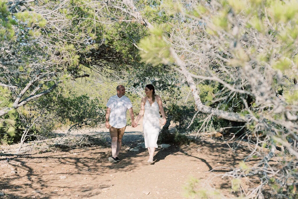 A couple walking hand-in-hand through a wooded area on a sunny day.