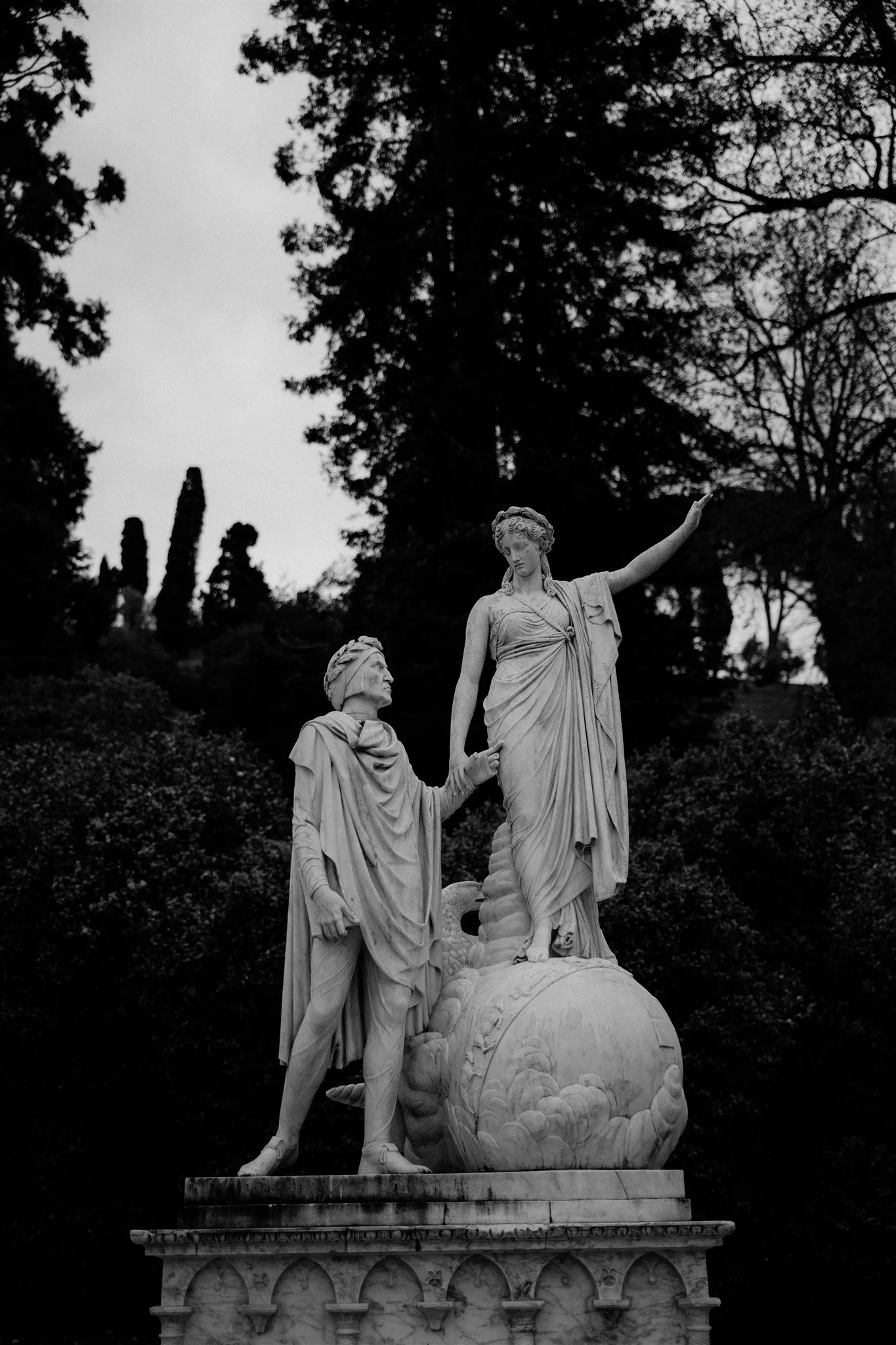 Black and white photograph of a classical marble statue featuring two figures, overlooking a detailed base with floral motifs, surrounded by trees and foliage.