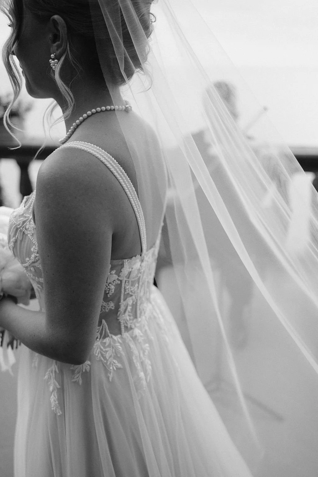 Black and white photo of a bride in a wedding dress with lace details, pearl necklace, pearl earrings, and a veil, standing sideways.