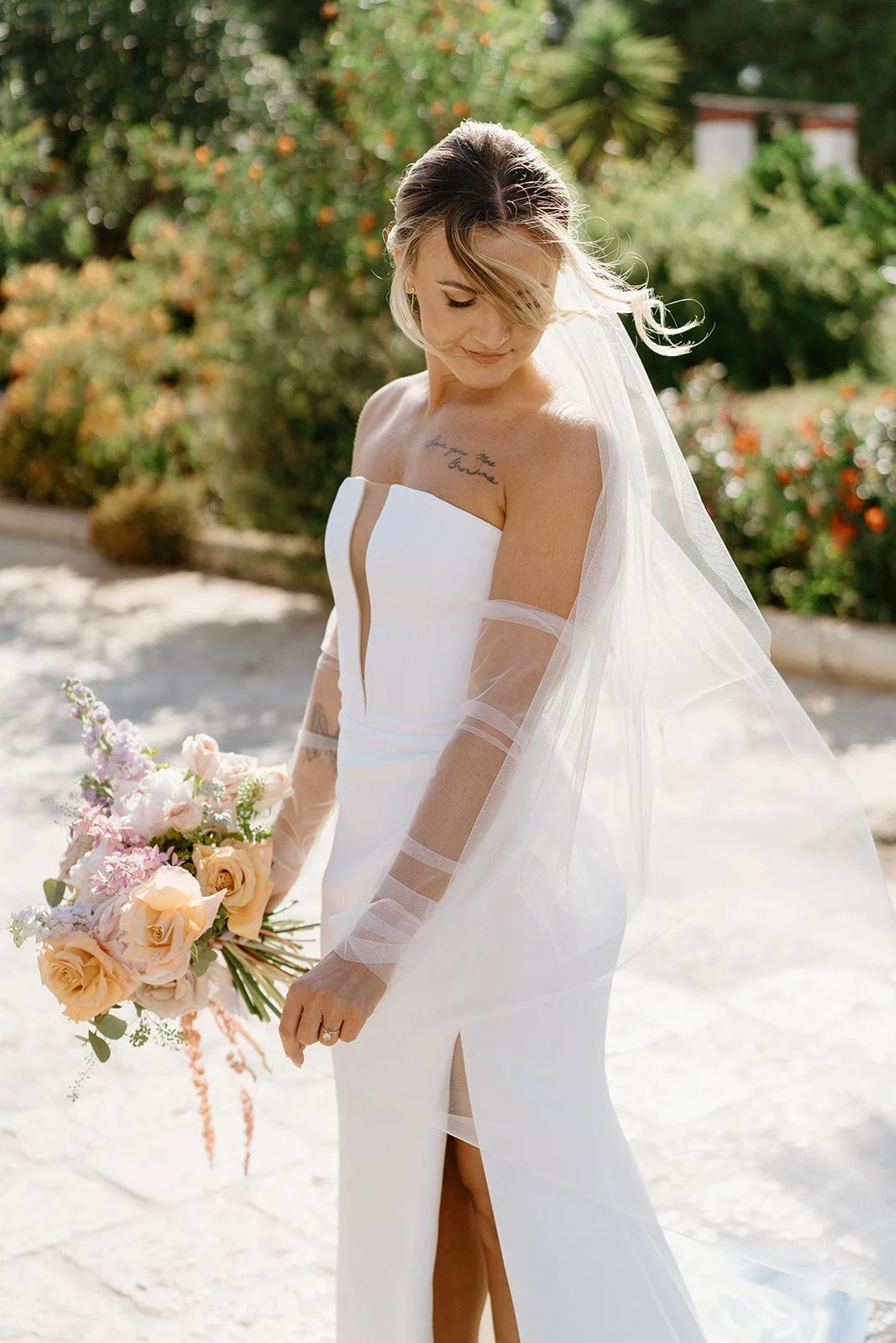A bride in a white strapless wedding gown with sheer sleeve accents, holding a bouquet of pink and peach roses, outdoors with green foliage and blooming flowers in the background.
