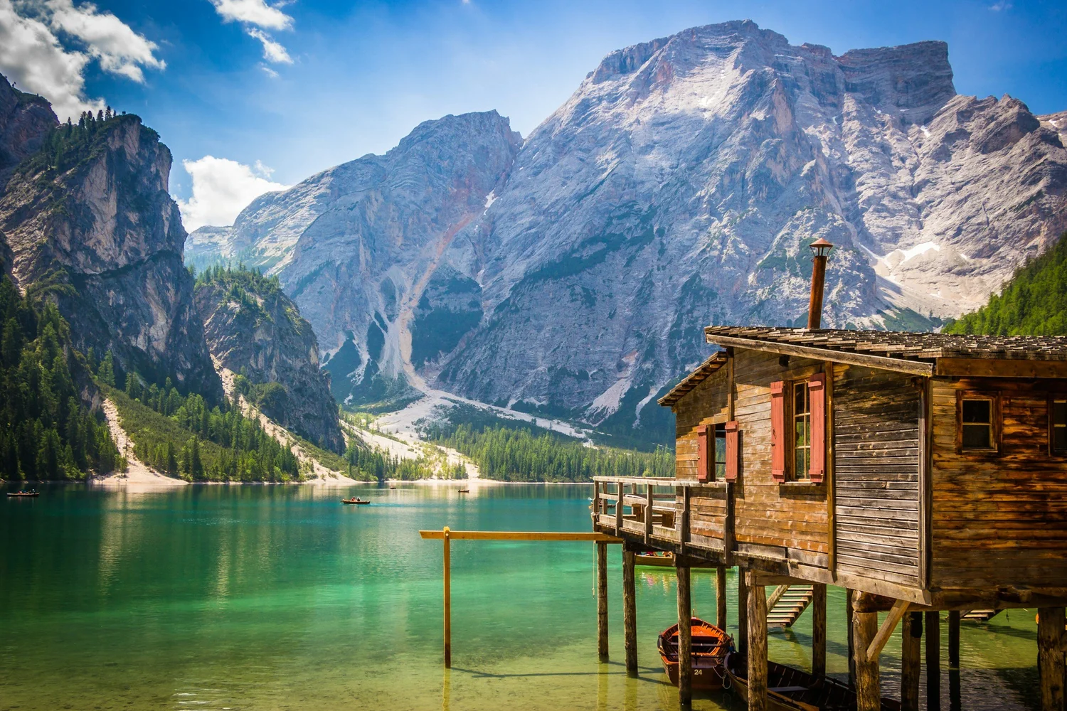 A wooden house built on stilts over a lake with turquoise water, surrounded by green trees and tall mountain cliffs in the background under a partly cloudy sky.