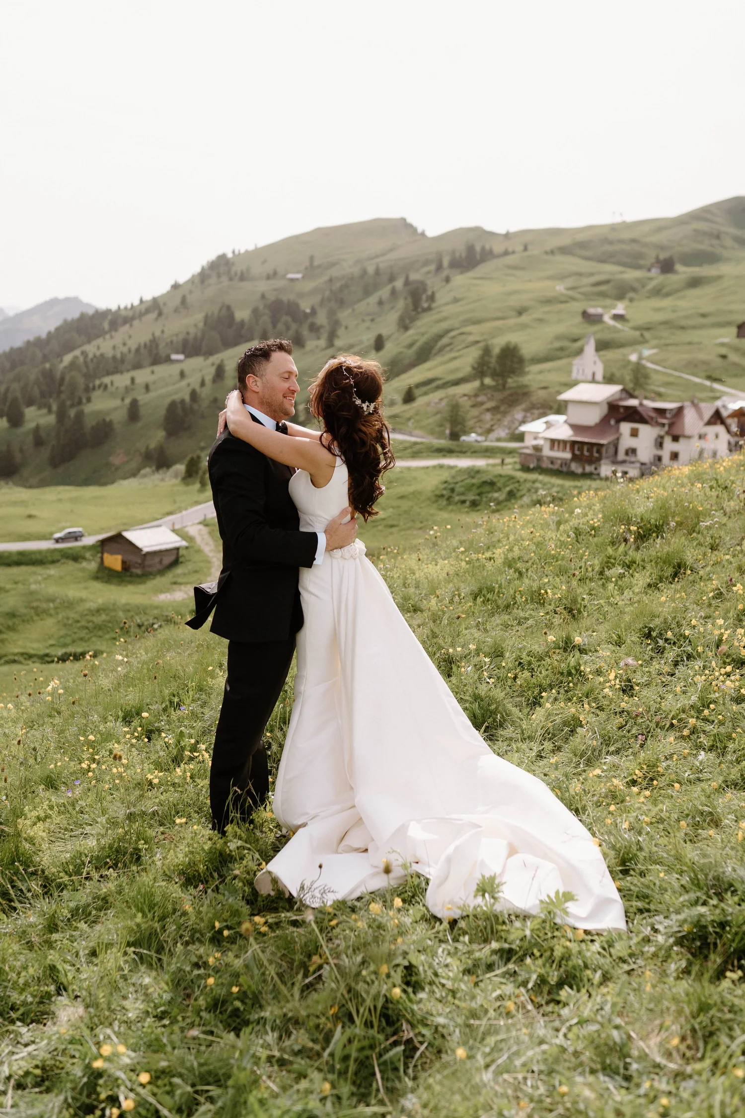 A newlywed couple dancing in a lush green meadow with yellow flowers, with rolling hills and scattered houses and a church in the background.