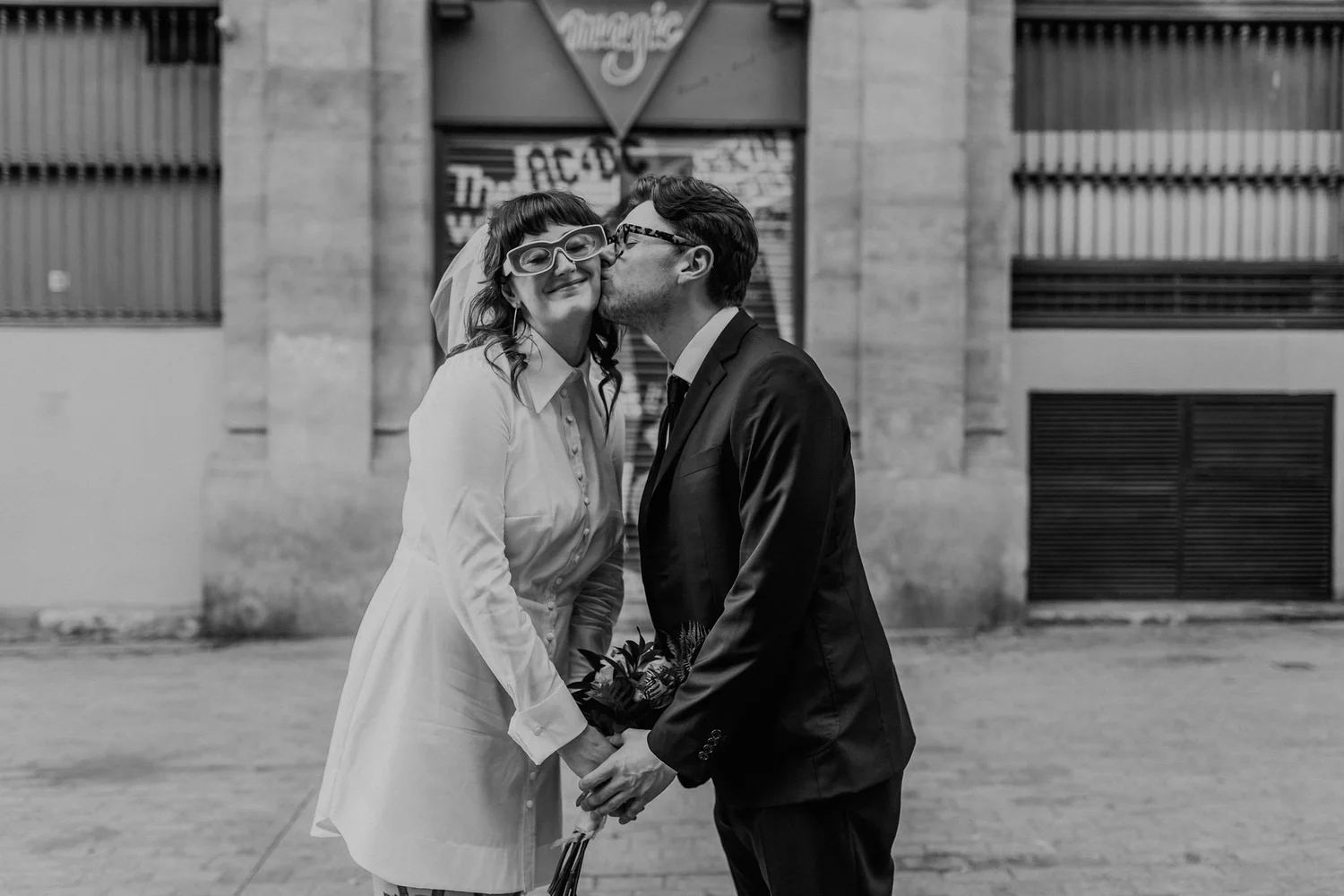 A couple dressed in wedding attire sharing a kiss outdoors, with the woman in a white dress and sunglasses, and the man in a suit and glasses, holding hands and a bouquet.