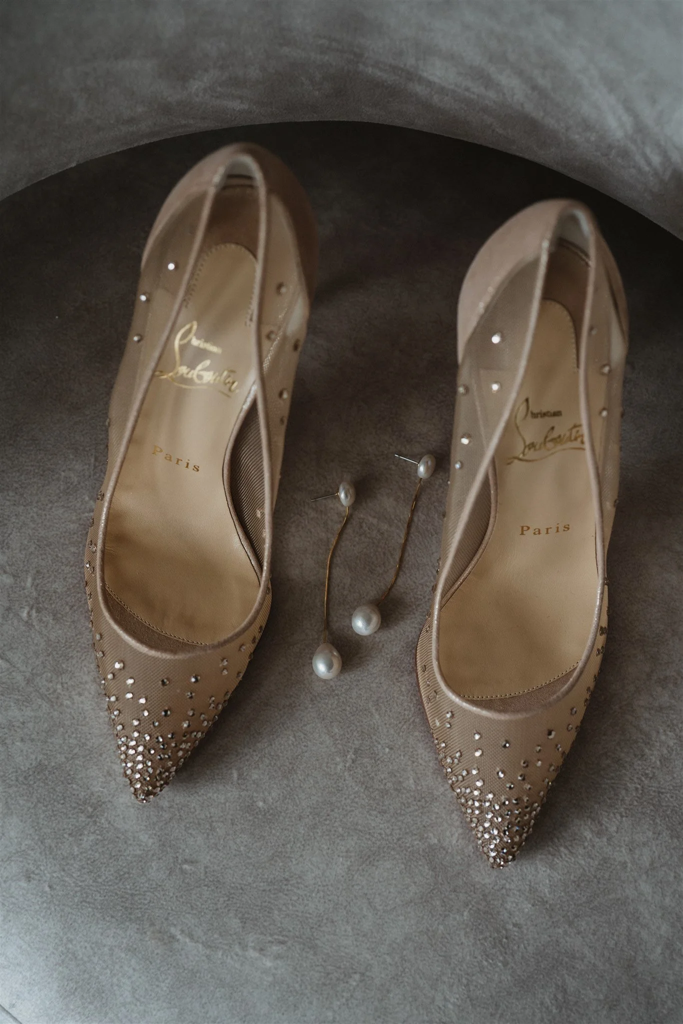 Beige pointed-toe high heels with embellishments and a pair of pearl earrings beside them, placed on a gray surface. The shoes have the name 'Christian Louboutin Paris' printed inside.