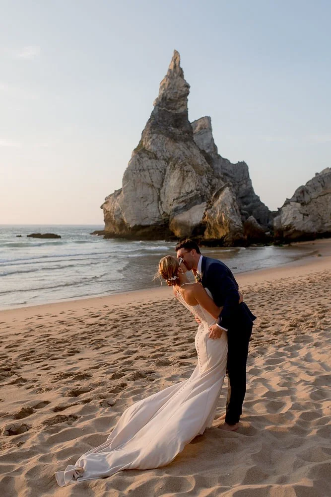 A bride and groom kissing on the beach with large rock formations in the background during sunset.