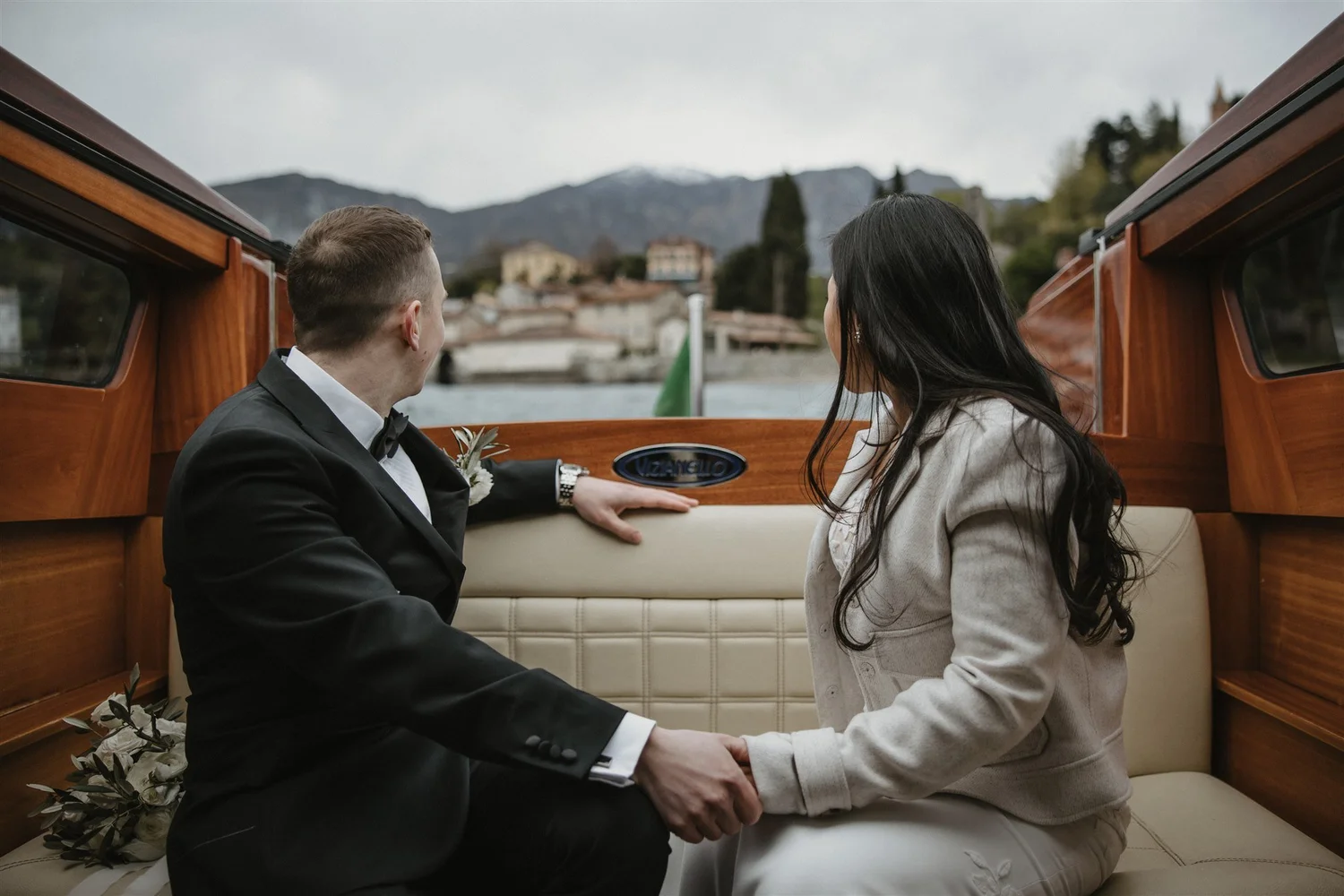 A groom and bride sitting inside a wooden boat, holding hands and facing each other, with a scenic mountainous town in the background.