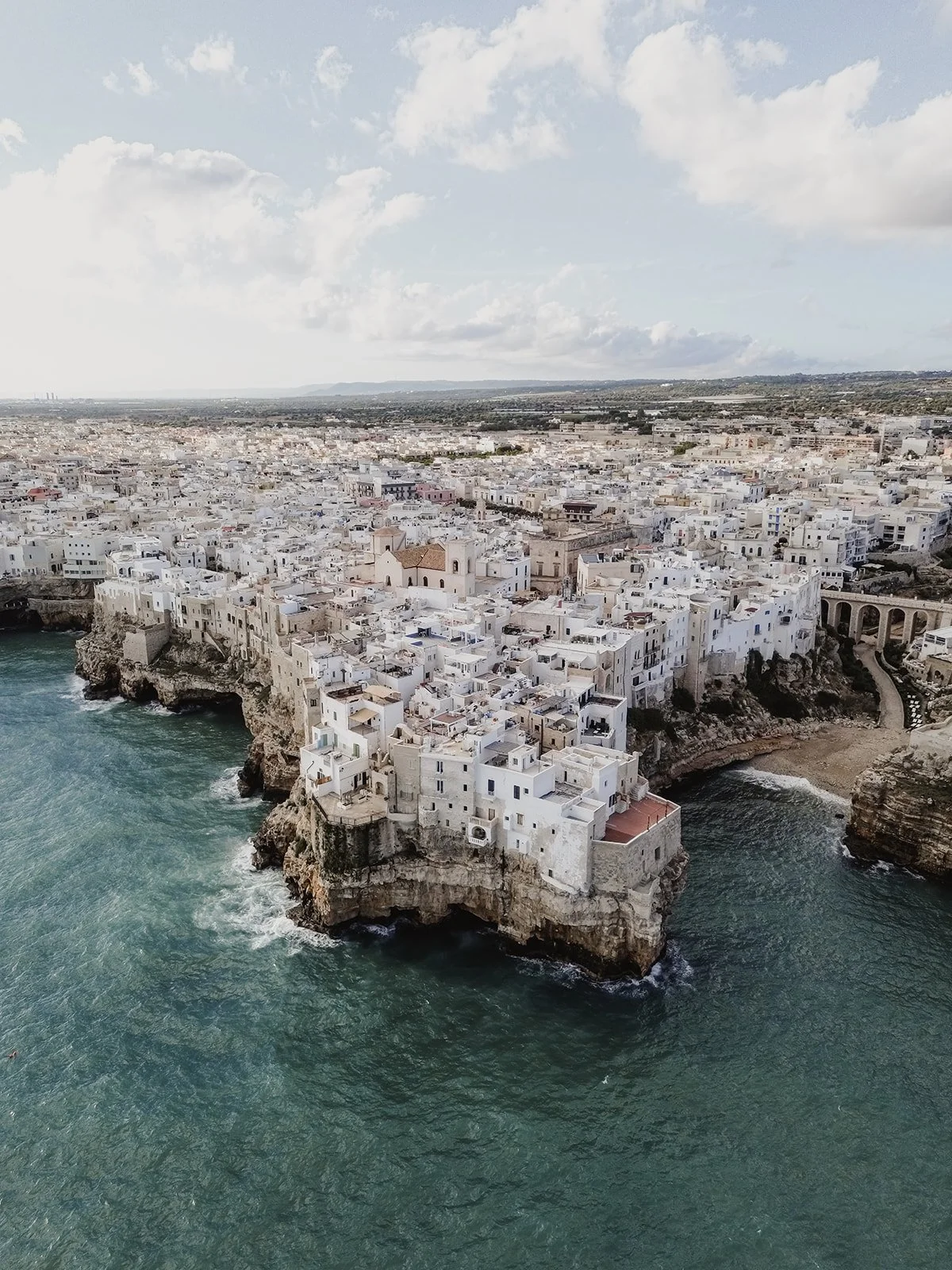 Aerial view of a coastal city with white buildings on cliffs along the ocean and a bridge in the background under a partly cloudy sky.