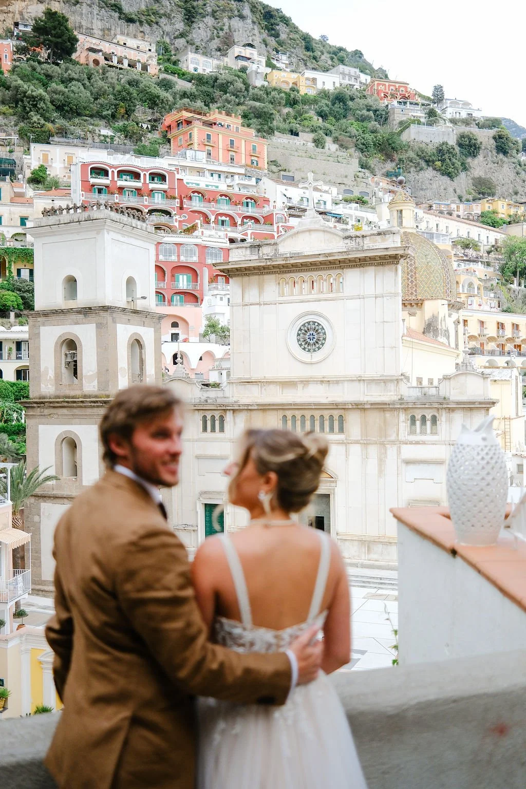 A couple in wedding attire smiling and embracing on a balcony with a scenic hillside town and a church in the background.