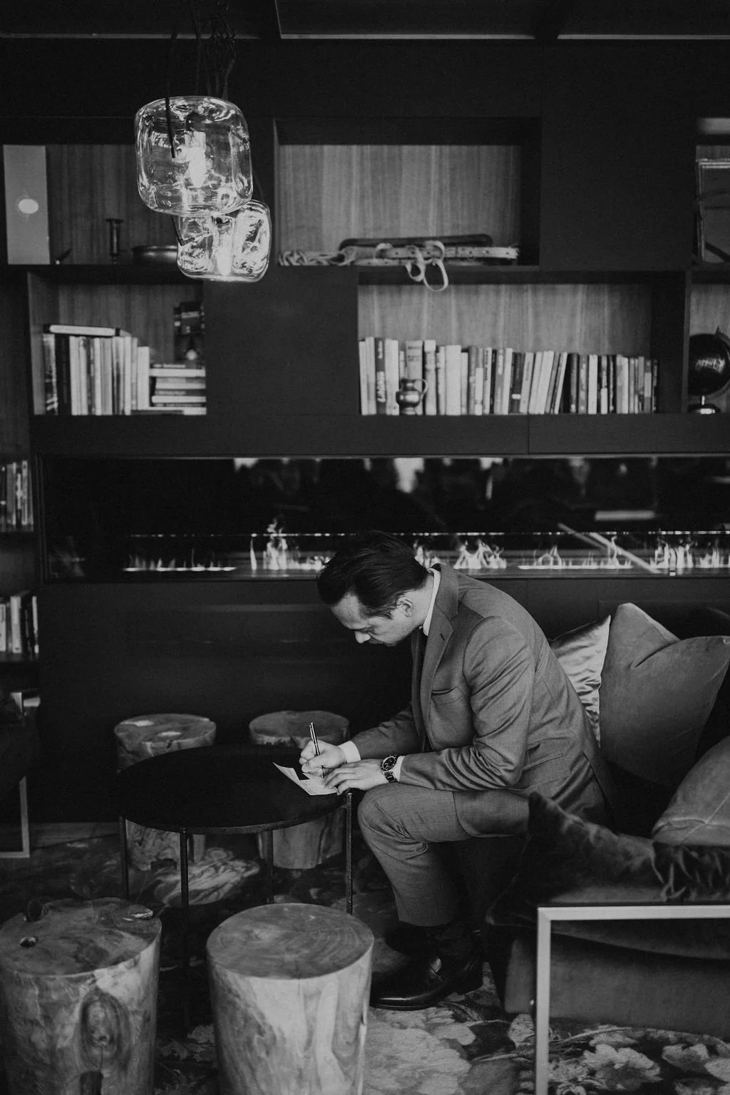 A man in a suit sitting on a sofa, writing on a piece of paper with a pen, in a room with bookshelves, a fireplace, and decorative elements.