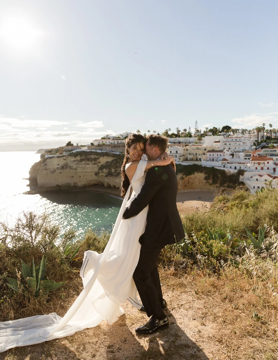 A newlywed couple embracing each other on a cliffside with a coastal town and ocean in the background.