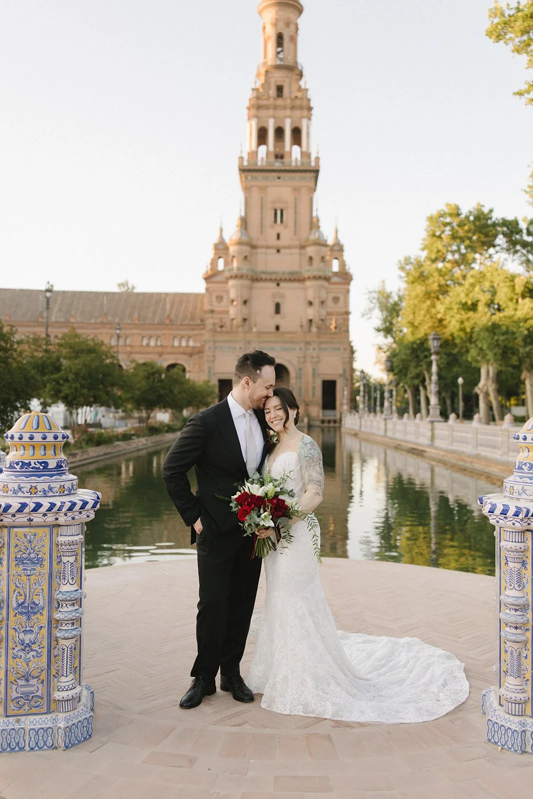 A wedding couple standing on a bridge over a waterway with a historic building in the background. The groom is in a black suit, and the bride in a white lace wedding dress holds a bouquet of red and white flowers. The scene is outdoors with lush trees and decorative ceramic columns on each side.