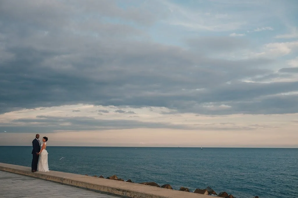 A couple dressed in wedding attire holding hands by the ocean at sunset.