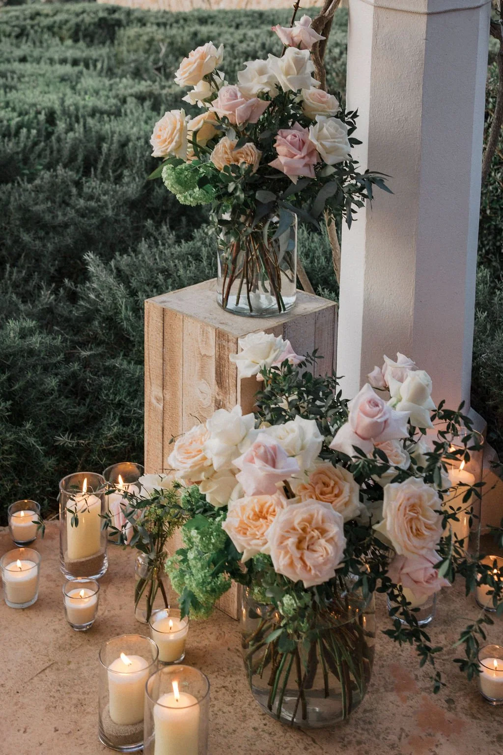 A floral arrangement of pink and white roses with greenery in glass vases on a table decorated with lit candles, set outdoors near a white wall and lush greenery.