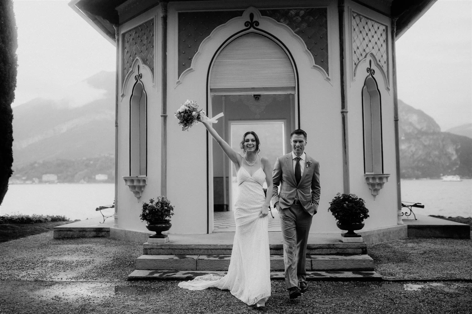 Black and white photo of a bride and groom standing hand in hand at the bottom of a short flight of stairs outside a decorative pavilion, with mountains in the background. The bride holds a bouquet in her raised hand and smiles.