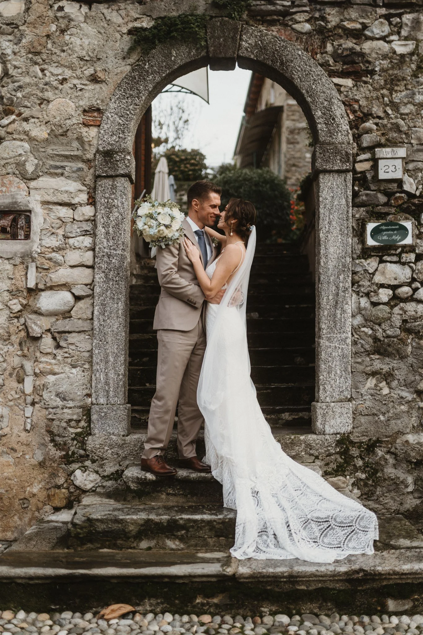 A bride and groom standing on stone steps beneath an arched stone doorway, embracing and smiling at each other during their wedding.