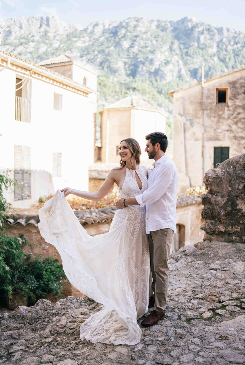 A happy couple, a woman in a white wedding dress and a man in a white shirt and beige pants, stands outdoors on a cobblestone path with rustic buildings and a mountain in the background, smiling at each other.