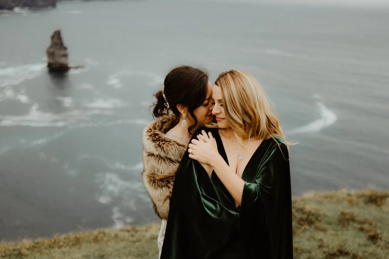 Two women hugging outdoors by a body of water, with a large rock or small island in the background, overcast weather, and fall foliage on the ground.