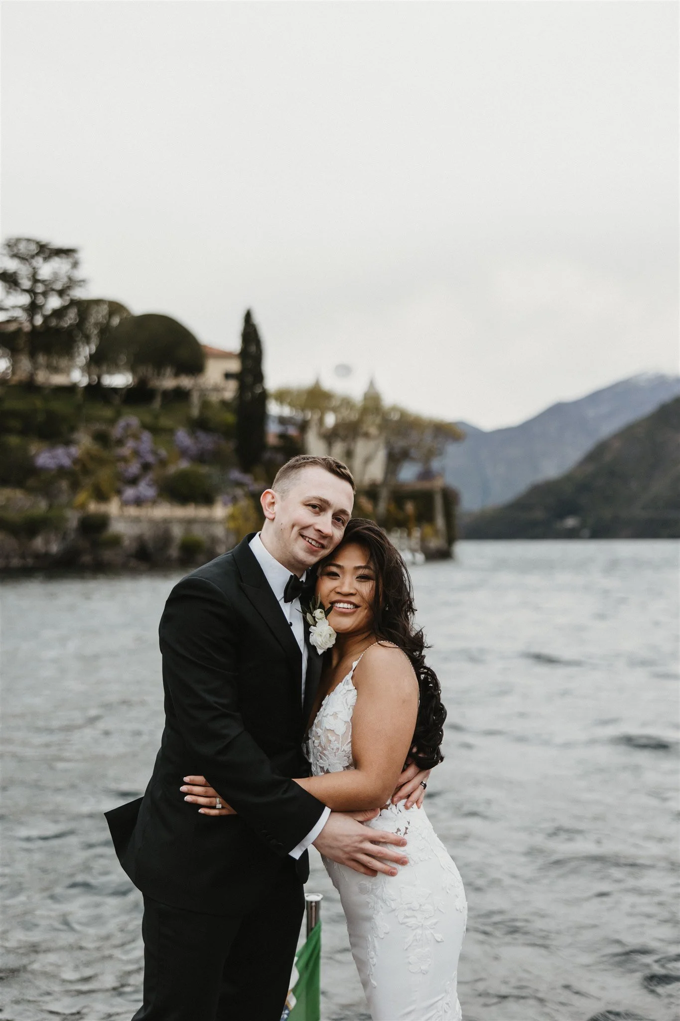 A newlywed couple smiling and embracing by a lake, with mountains in the background and trees on the hillside.