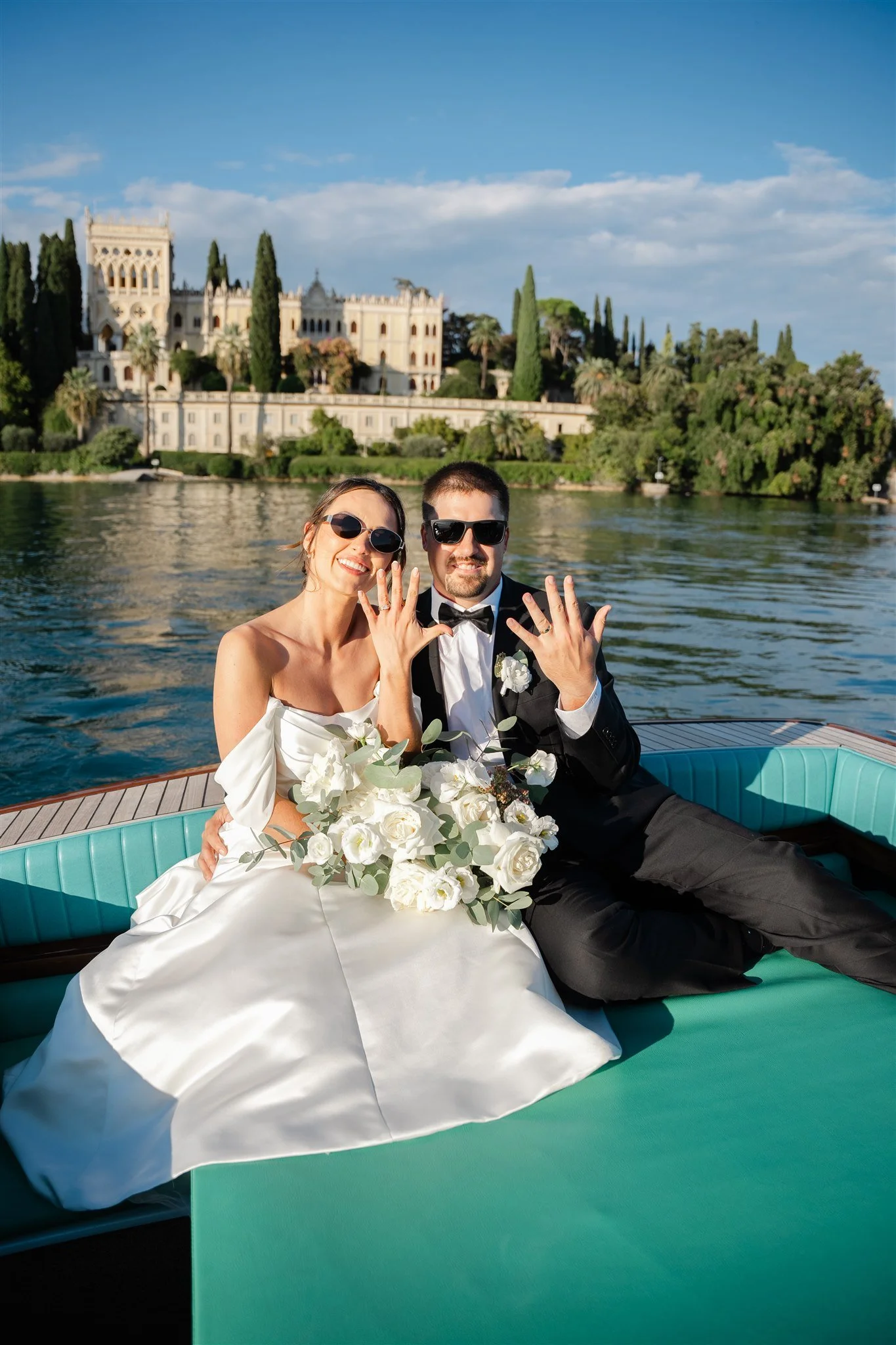 A newlywed couple dressed in wedding attire, sitting on a boat on a river, showing off their wedding rings and smiling. The bride is holding a bouquet of white flowers, and both are wearing sunglasses. In the background, there are lush trees and an elegant historic building.