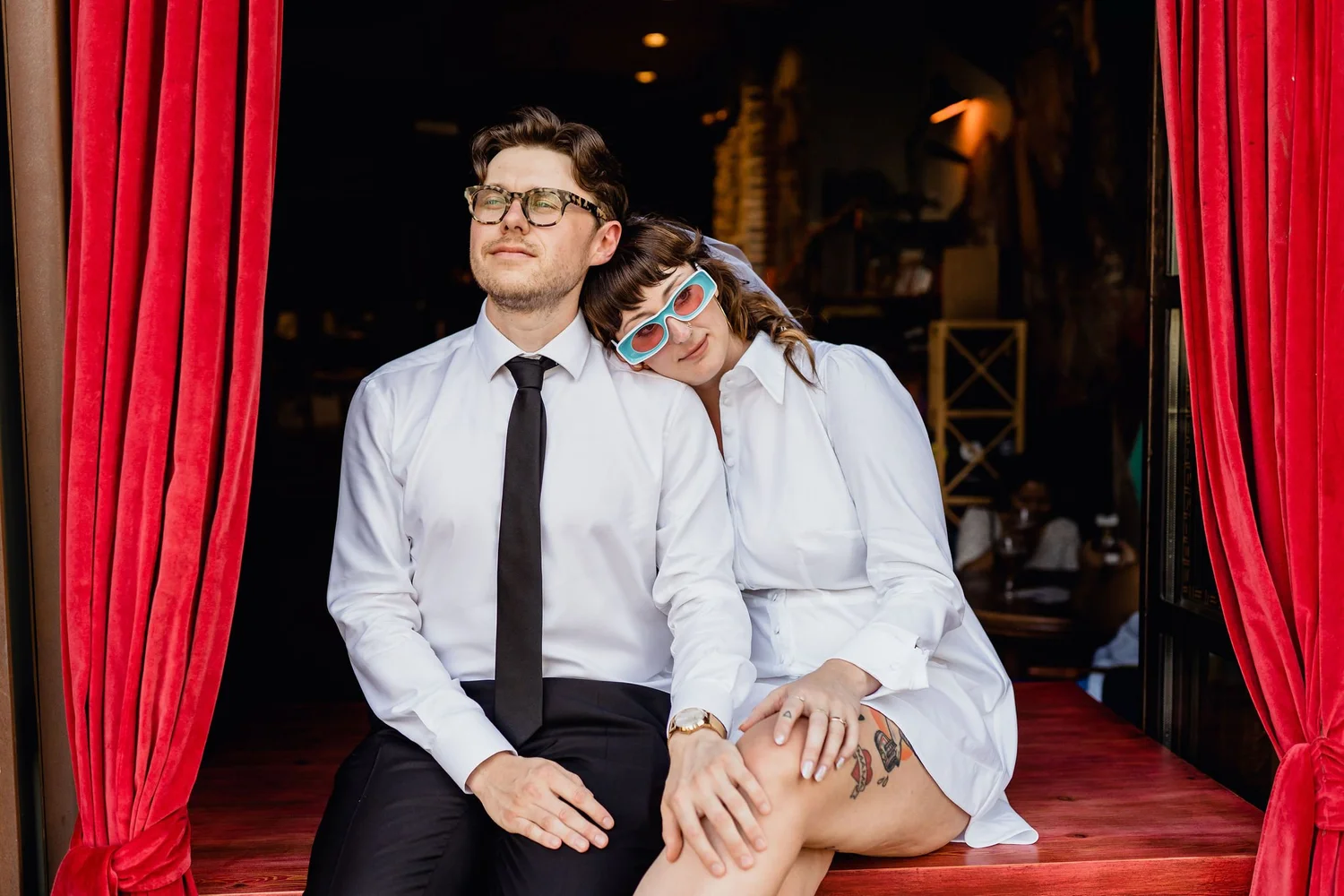 A man and woman sitting on a stage with red curtains. The woman is resting her head on the man's shoulder, both wearing white shirts and sunglasses. The man has glasses, dark hair, and a black tie; the woman has short brown hair with bangs and is wea