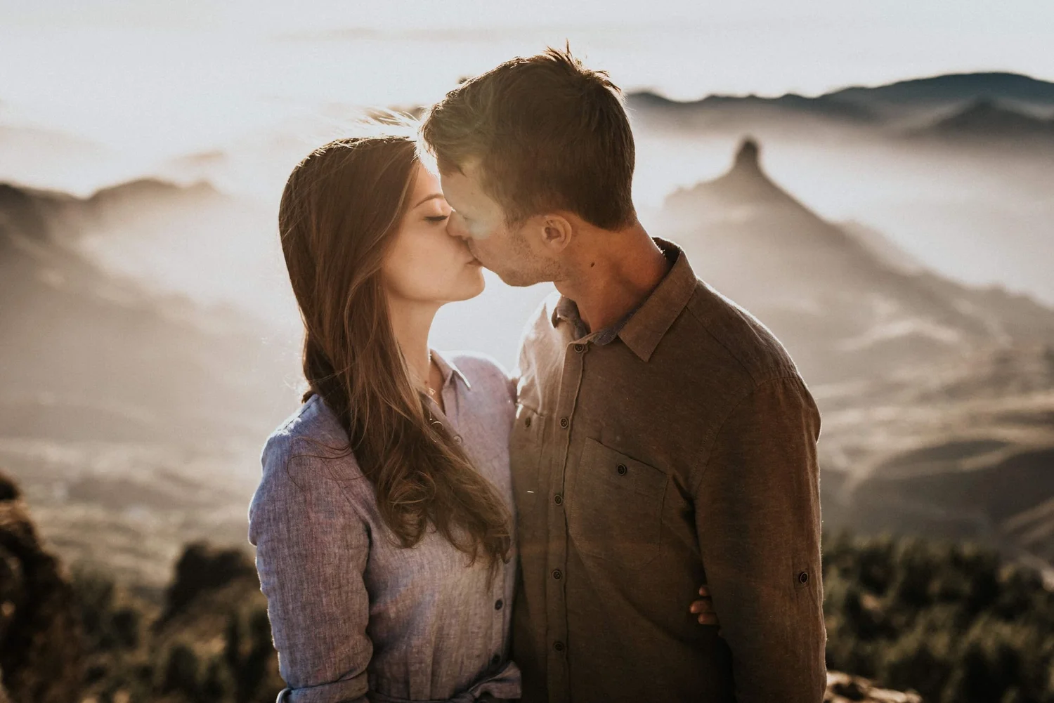 A couple sharing a kiss on a beach at sunset, with mountains in the background.