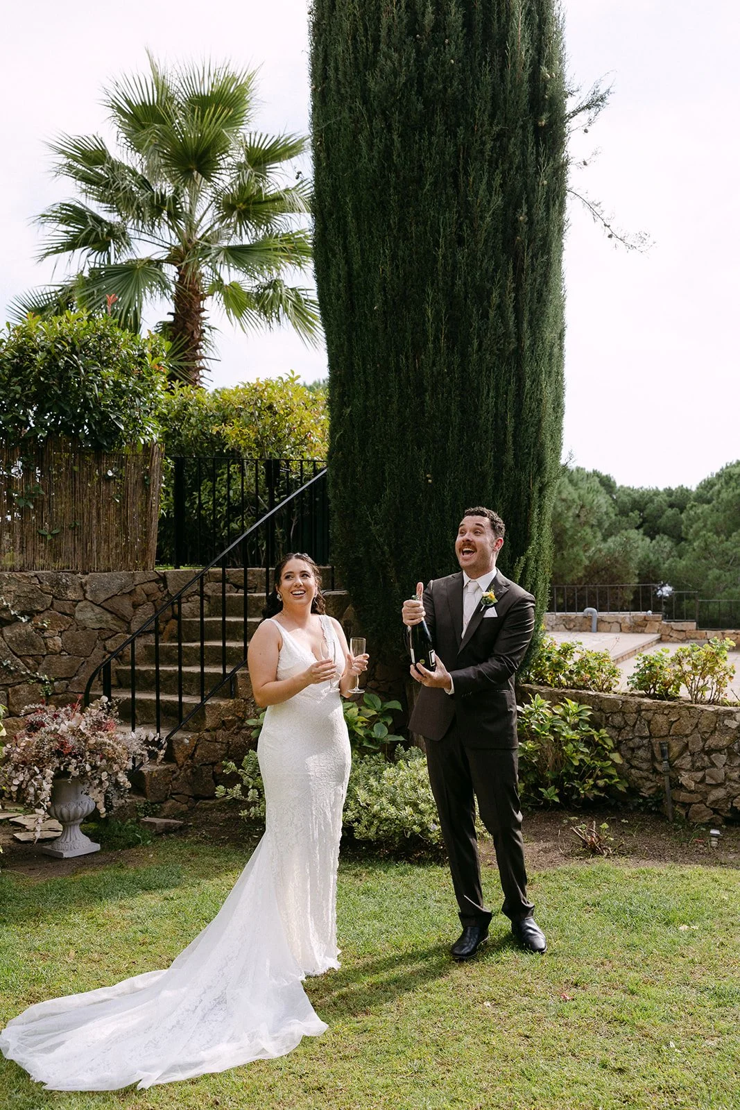 Bride and groom celebrating outdoors, with the bride in a white wedding dress holding a glass of champagne and the groom in a black tuxedo opening a bottle of champagne, surrounded by greenery and stone steps.
