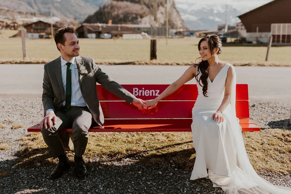 A bride and groom sitting on a red bench outdoors, holding hands and smiling at each other. The groom is wearing a gray suit with a white shirt and green tie, while the bride is in a white wedding dress with long dark hair. The background features a
