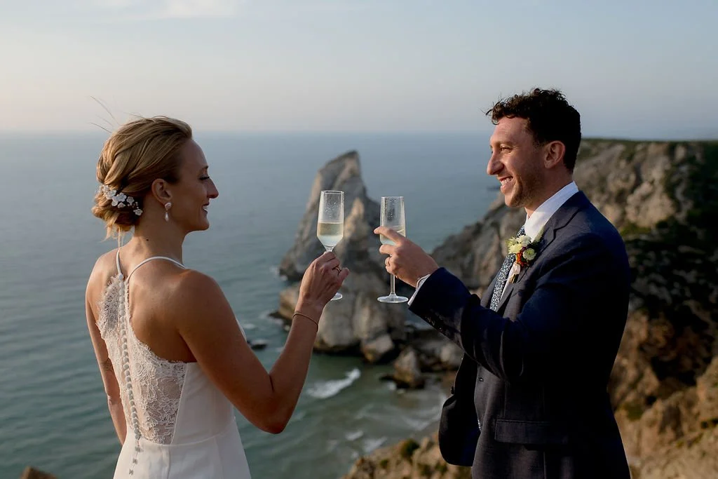 A bride and groom celebrating with champagne at a scenic coastal cliff scene during their wedding.