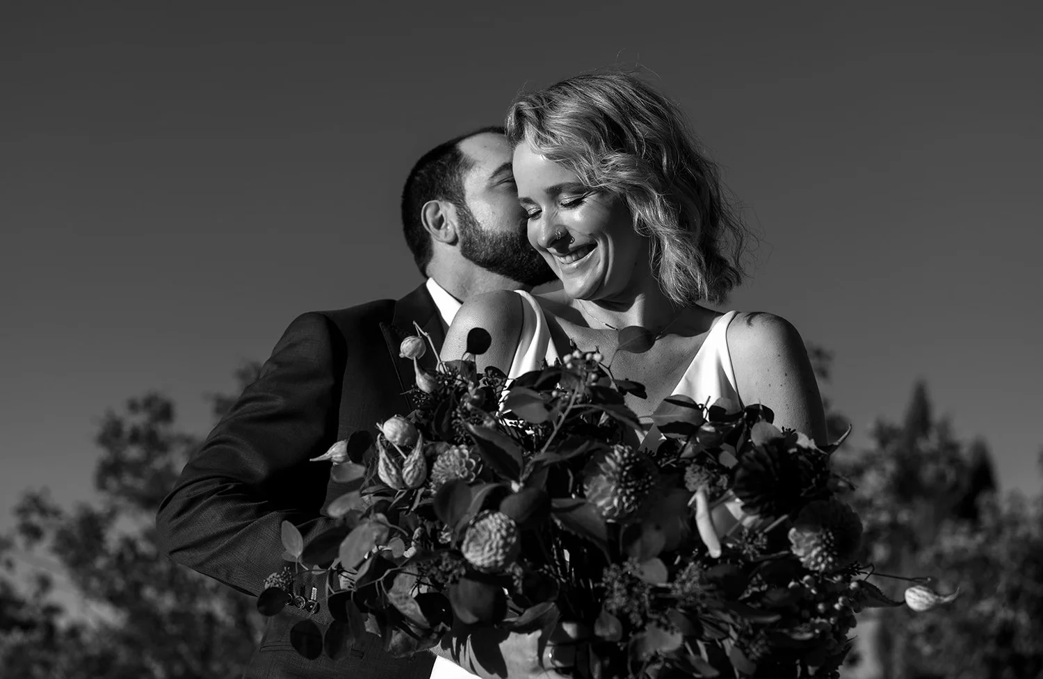 A black-and-white photo of a couple at their wedding, with the groom kissing the bride on the cheek while holding a bouquet of flowers. The bride is smiling with her eyes closed, wearing a dress and a floral necklace, and the groom is in a suit. The