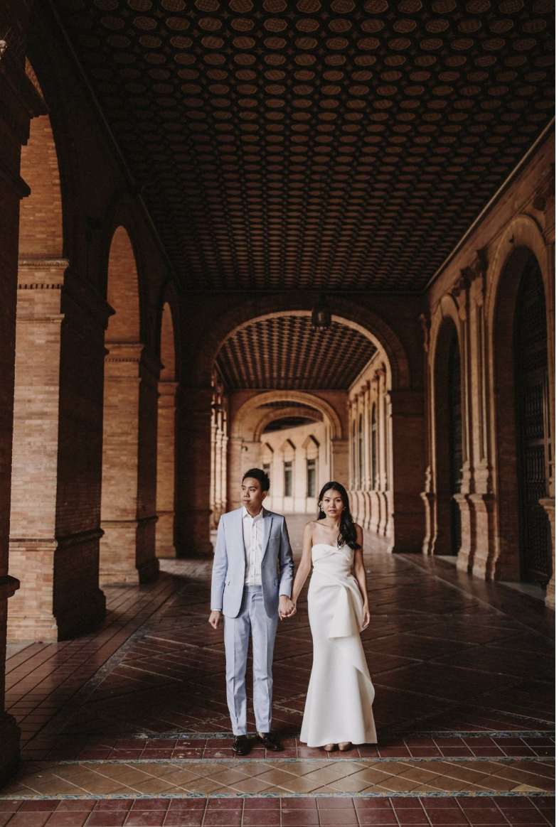 A couple dressed in formal wedding attire walking hand in hand through a covered, brick architectural corridor.