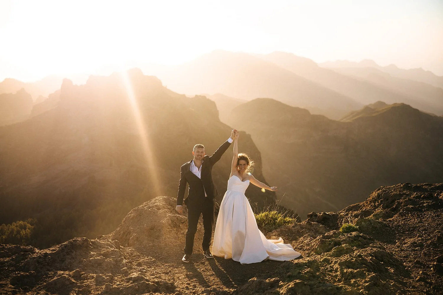 A newlywed couple in wedding attire standing on a rocky mountain with a mountain range in the background during sunset, celebrating with a raised hand.