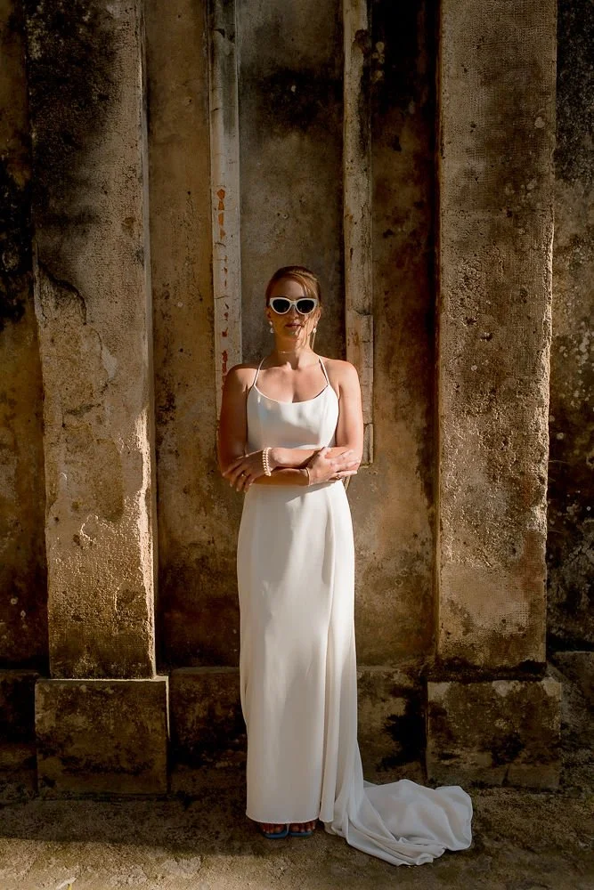 Woman in a white dress and sunglasses standing against a rustic stone wall.