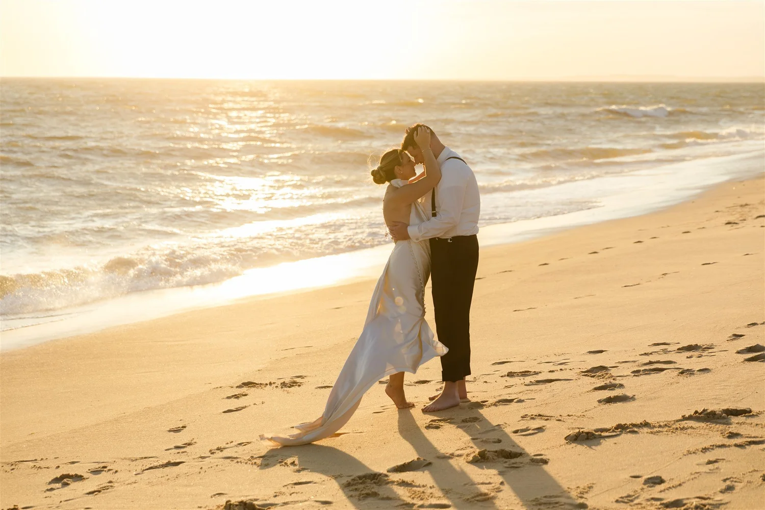A couple dressed in wedding attire, embracing and touching foreheads on a sandy beach during sunset.