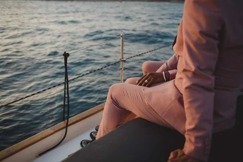 A person sitting on a boat near the water, wearing a pink outfit and a bracelet, looking out at the ocean during sunset.