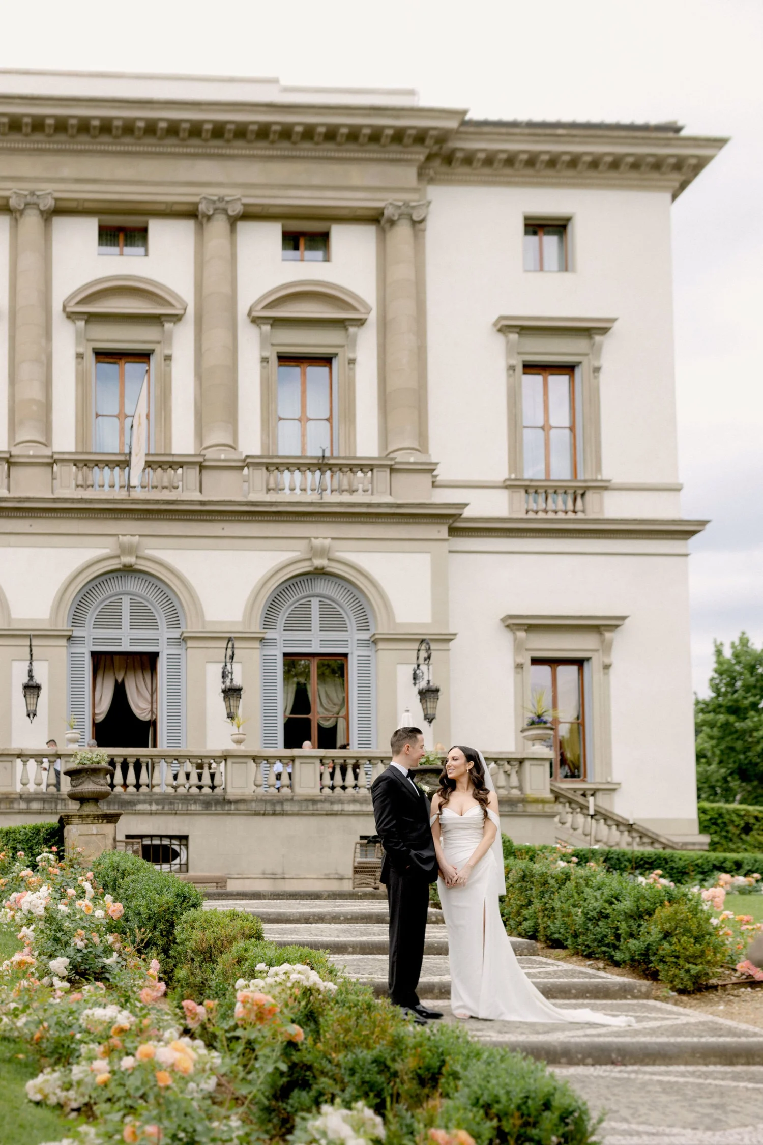 A bride and groom in formal wedding attire standing outdoors on stone steps in front of a large, elegant mansion with multiple windows and ornate architectural details, surrounded by garden flowers.
