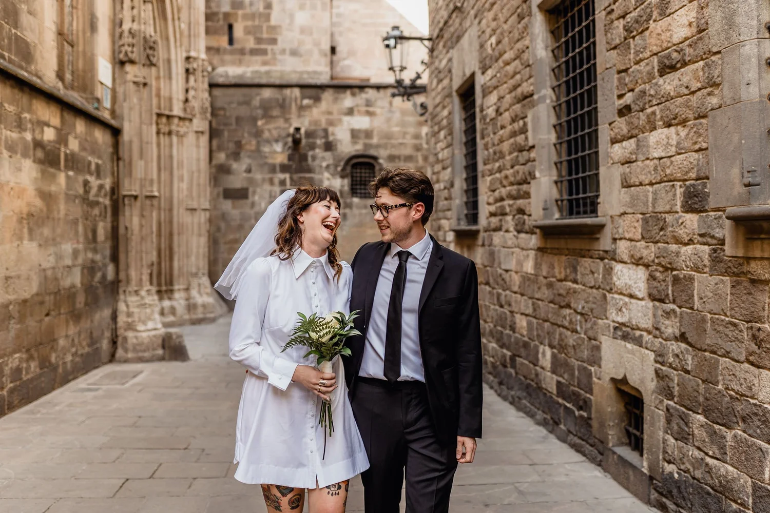 A groom and bride walking together in a narrow street with stone walls. The bride is holding a bouquet of greenery, and both are smiling happily. The bride is wearing a white dress and a veil, while the groom is dressed in a black suit with a tie.