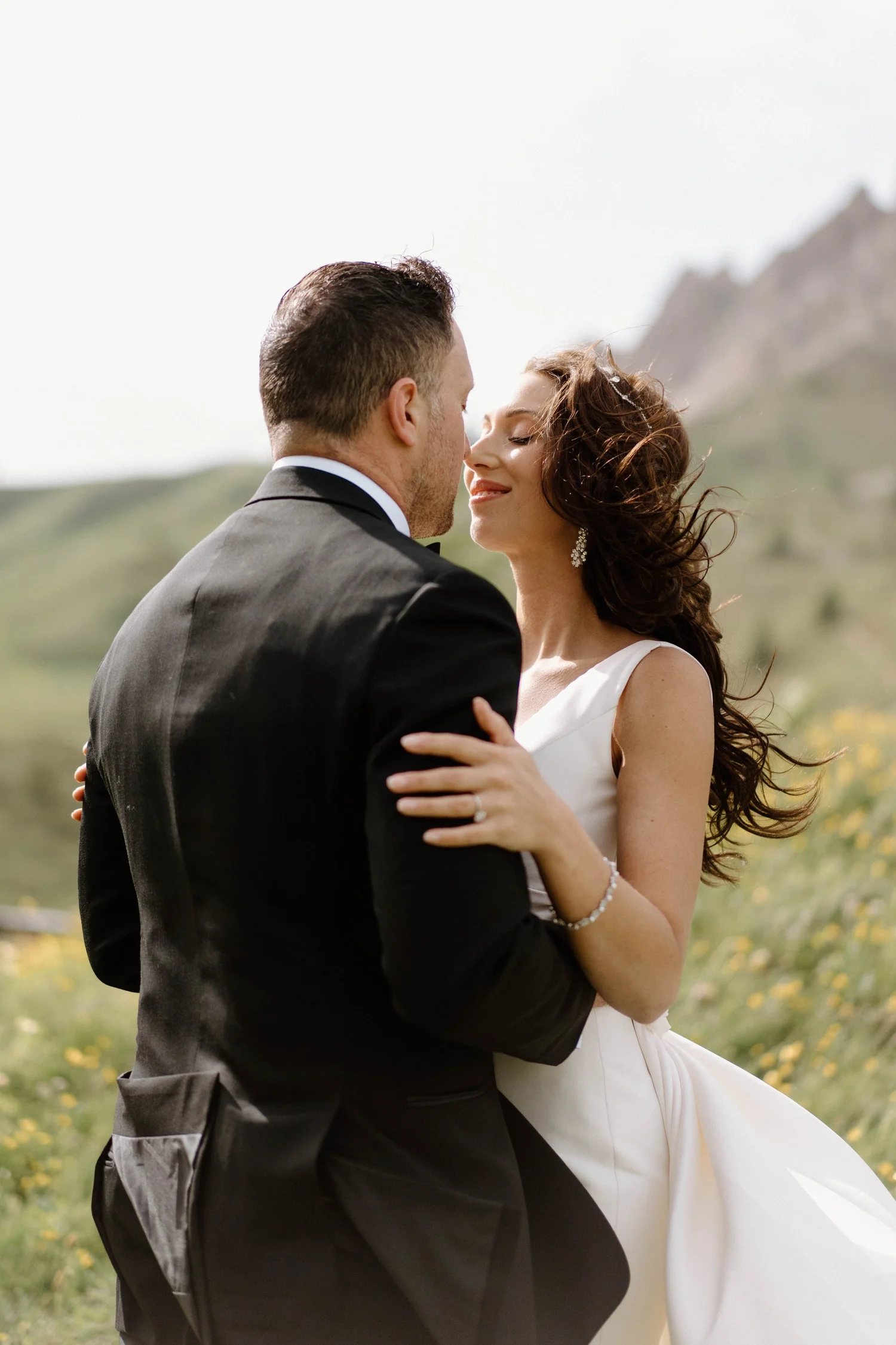 A bride and groom sharing a kiss outdoors in a scenic, mountainous area with greenery and yellow wildflowers.