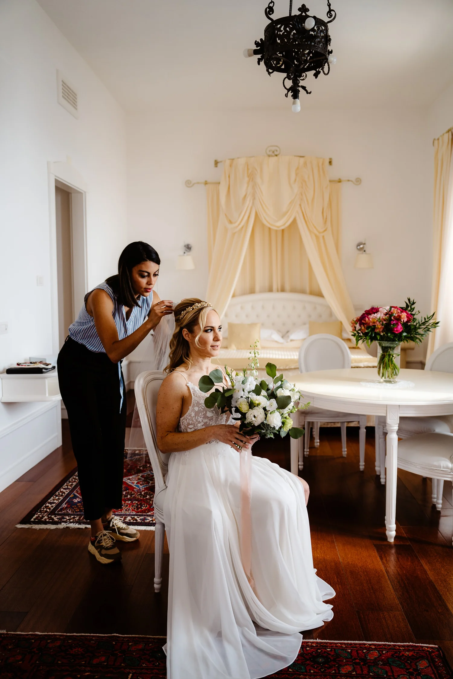 A bride sitting in a white chair holding a bouquet of white flowers, as a woman in a striped top styles her hair in a bright, elegant bedroom with a four-poster bed with cream curtains, a bouquet on the table, and a black chandelier overhead.