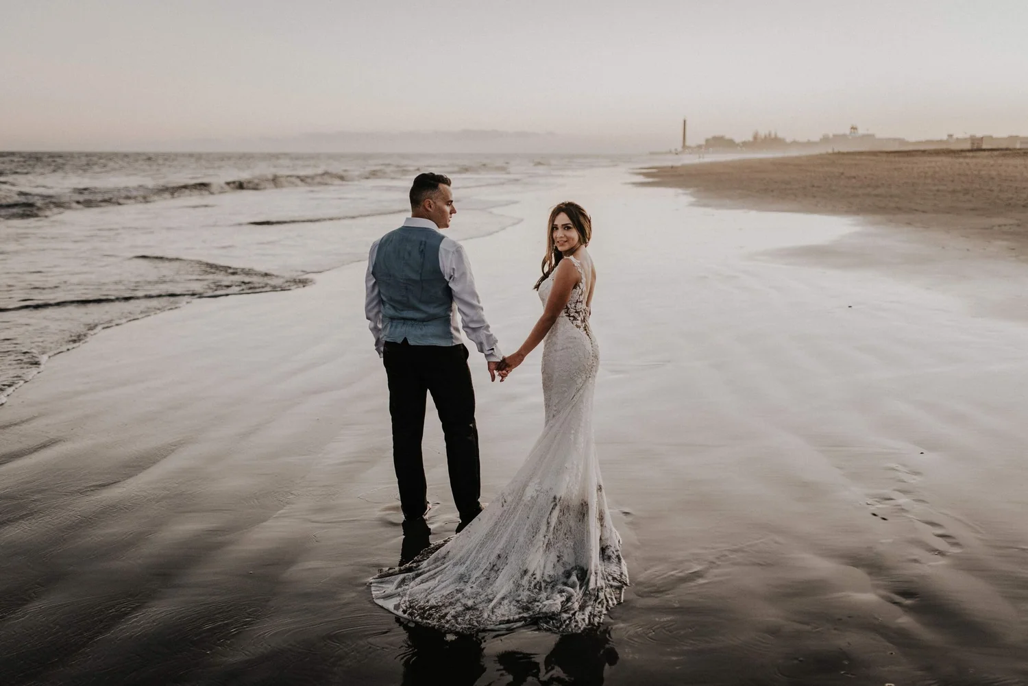 A couple in wedding attire holding hands and walking in shallow ocean water at sunset on the beach, with distant shoreline and buildings in the background.