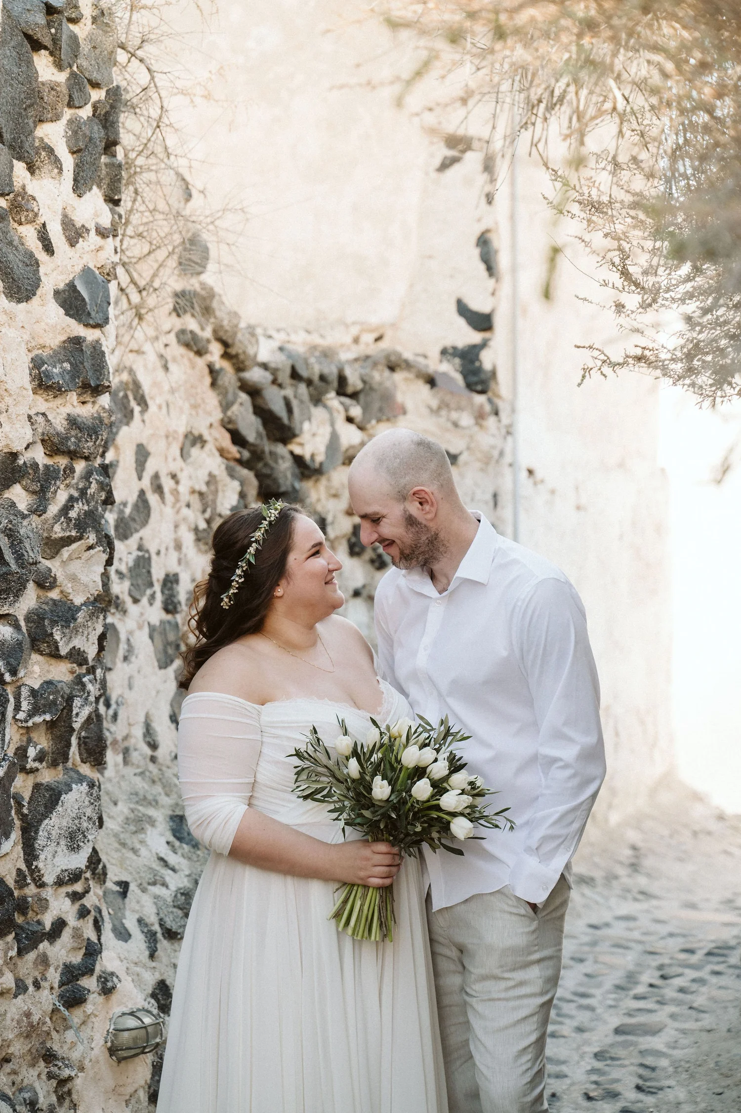A bride and groom standing close together outdoors, smiling and looking at each other. The bride is holding a bouquet of white tulips and wearing a white dress and floral headband. The groom is dressed in a white shirt and light-colored pants, with a
