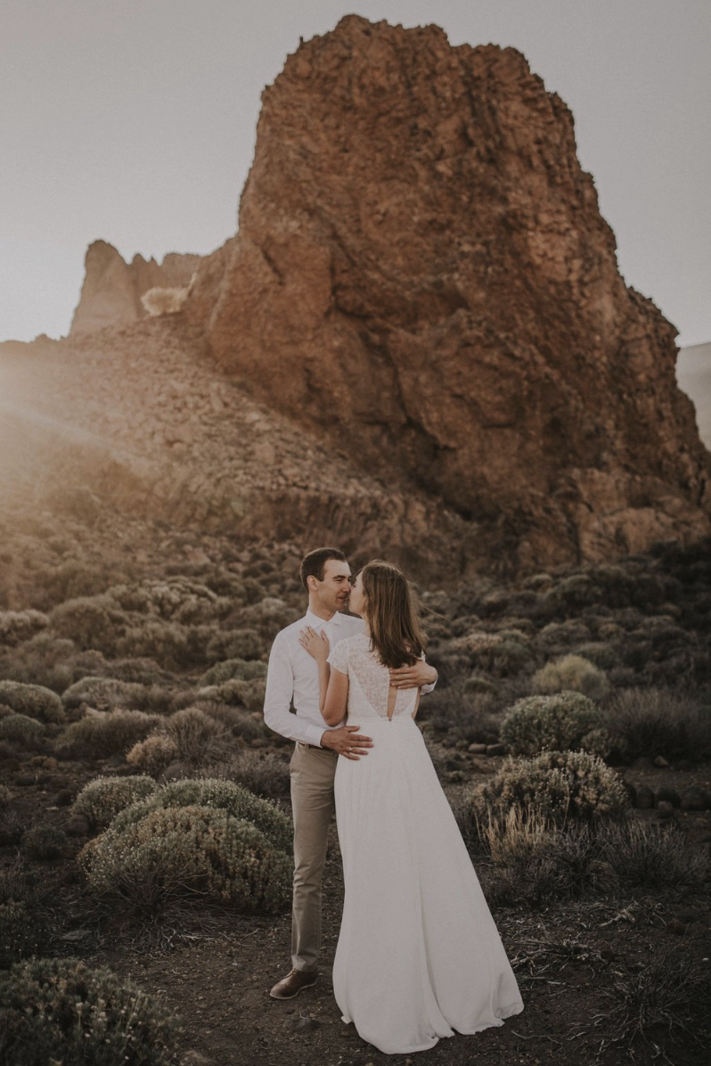 A couple in wedding attire standing in a desert landscape with large rock formations and shrubbery, embracing each other at sunset.