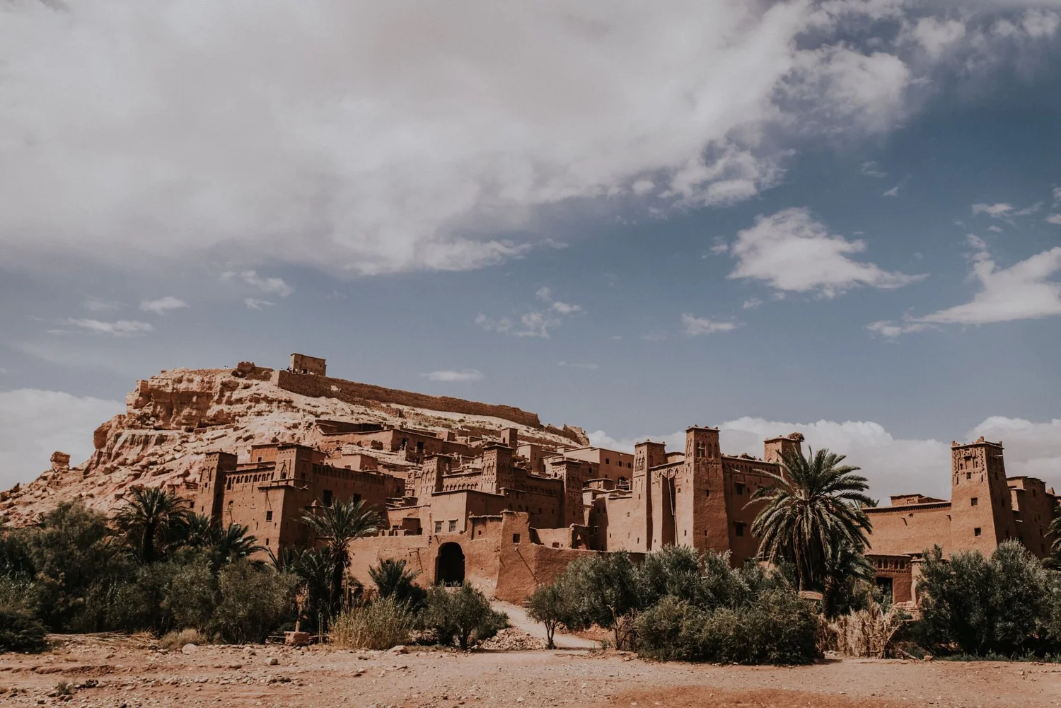 Ancient mud brick kasbah complex on a hill with palm trees in front, under a partly cloudy sky.