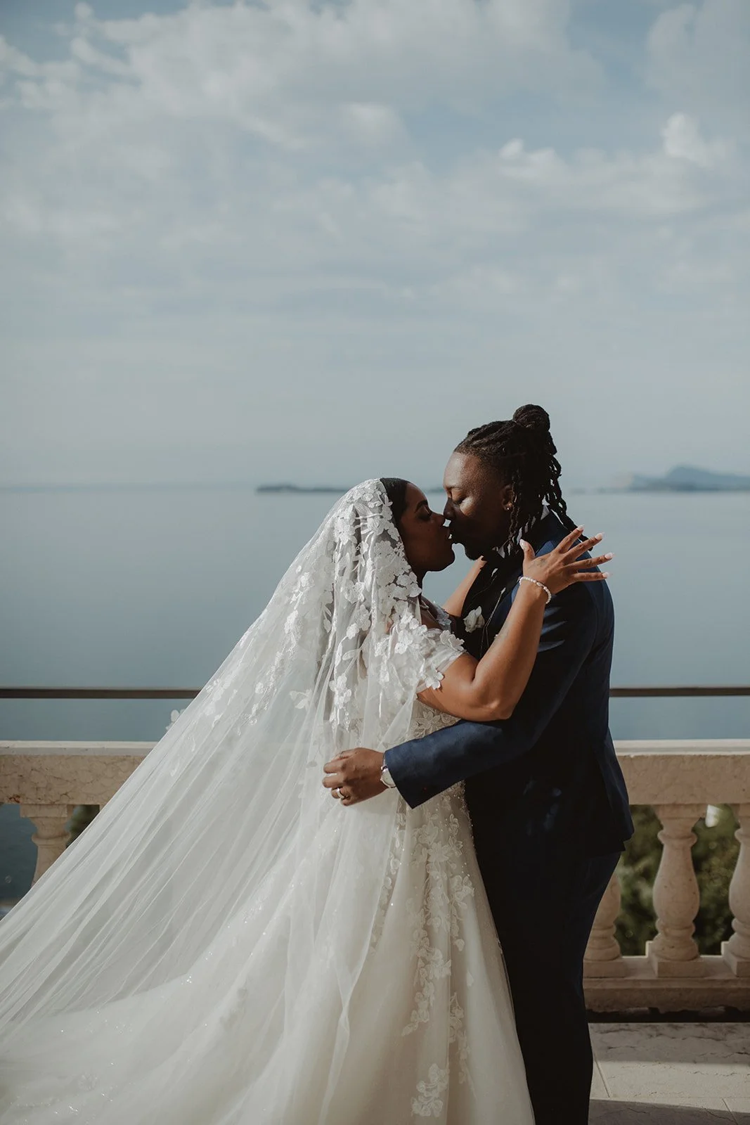 A bride and groom sharing a kiss on a balcony overlooking a body of water, with cloudy sky above.