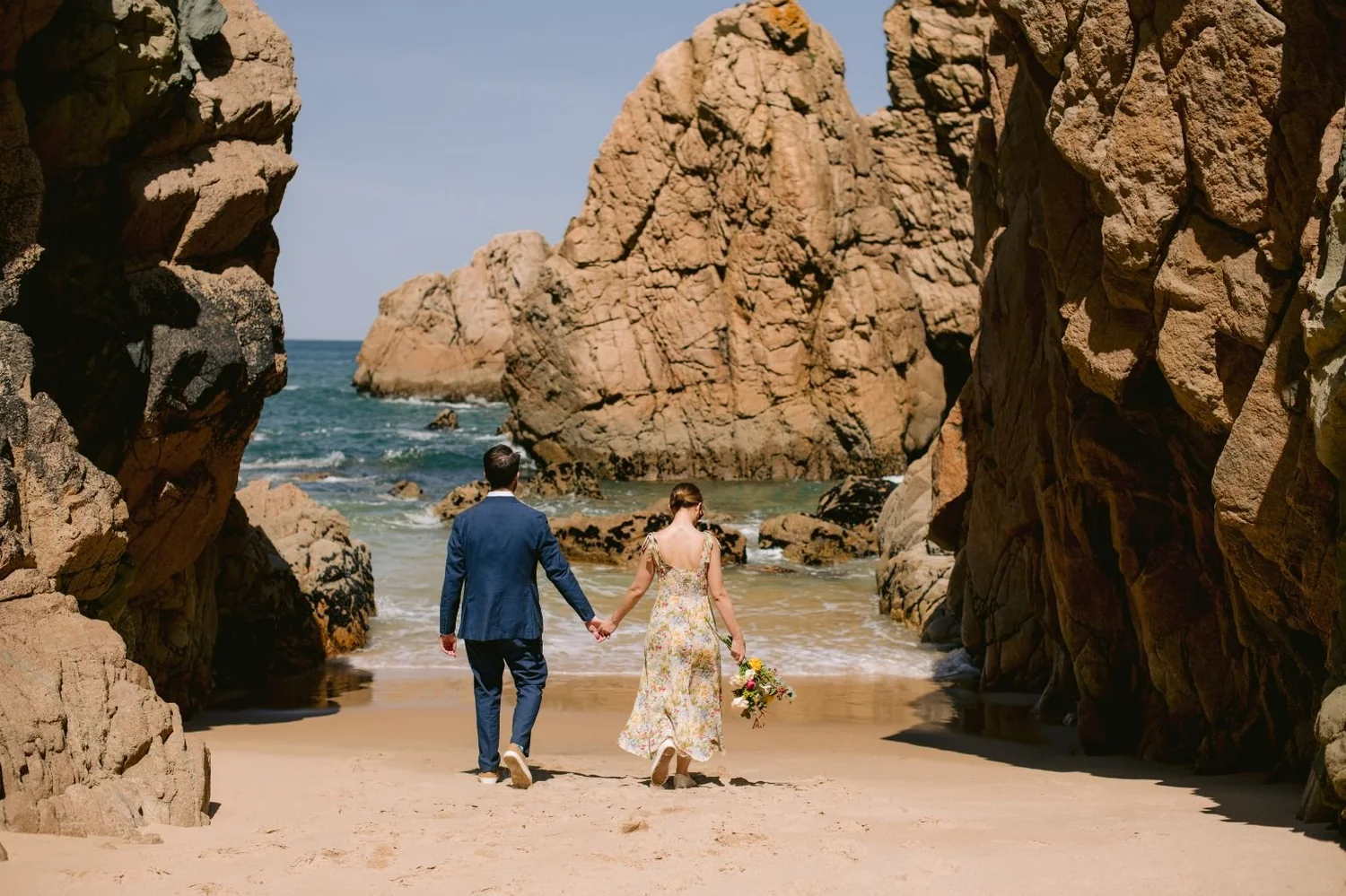 A couple walking hand in hand on a sandy beach between large rock formations, with the ocean in the background. The woman is holding a bouquet of flowers.