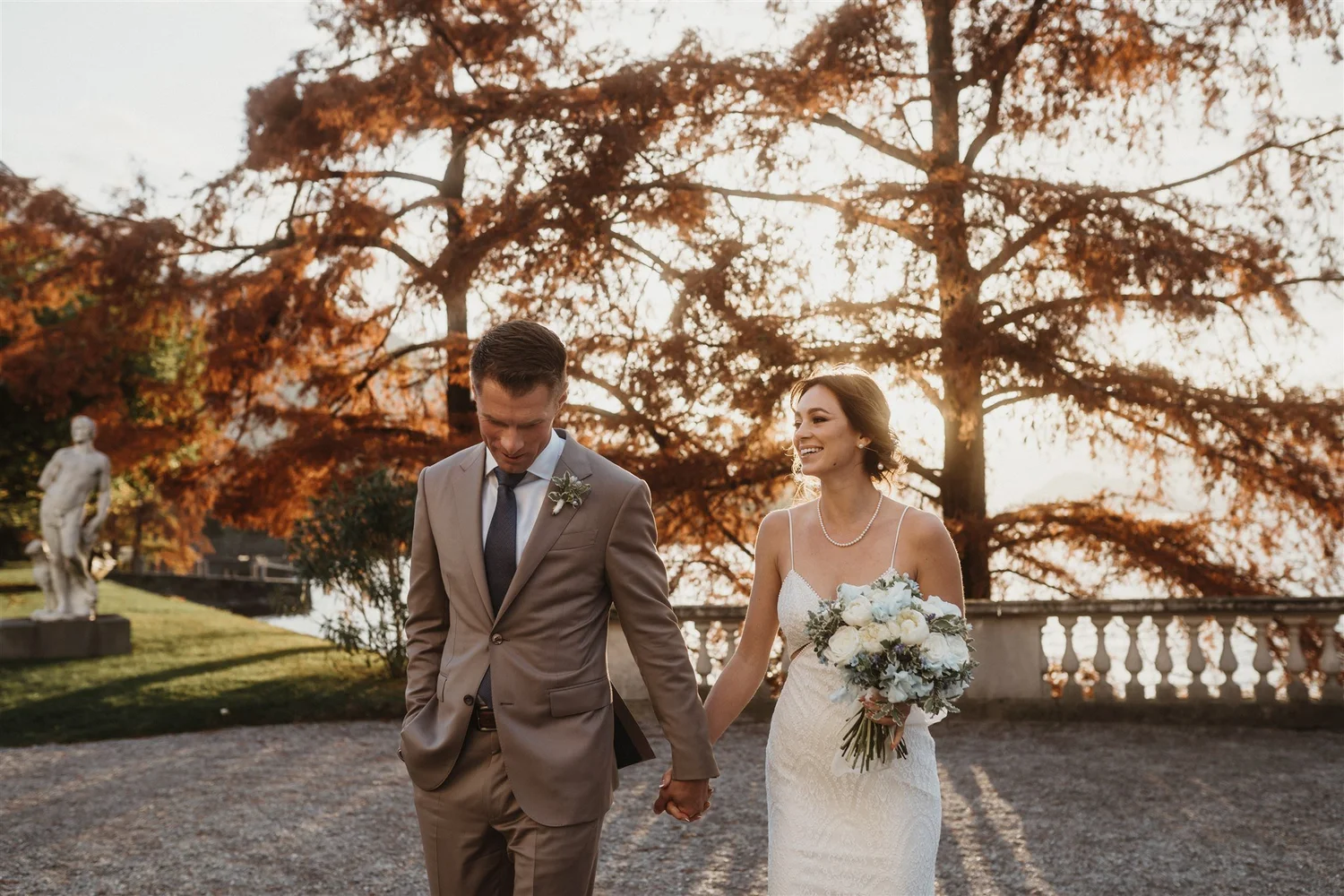 A bride and groom walking hand in hand outdoors during a wedding, with autumn leaves and trees in the background.
