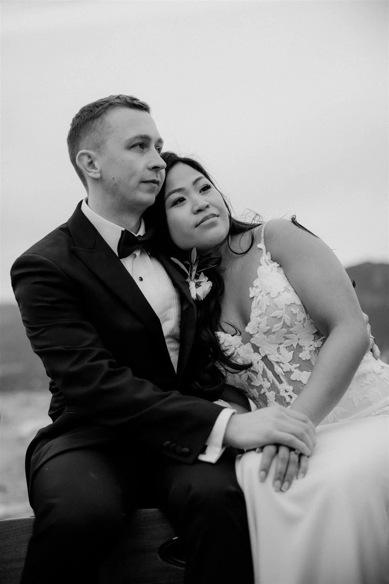 A black-and-white photograph of a married couple sitting closely together outdoors. The groom wears a tuxedo with a bow tie, and the bride wears a lace wedding dress. They are holding hands, and the bride rests her head on the groom's shoulder, both